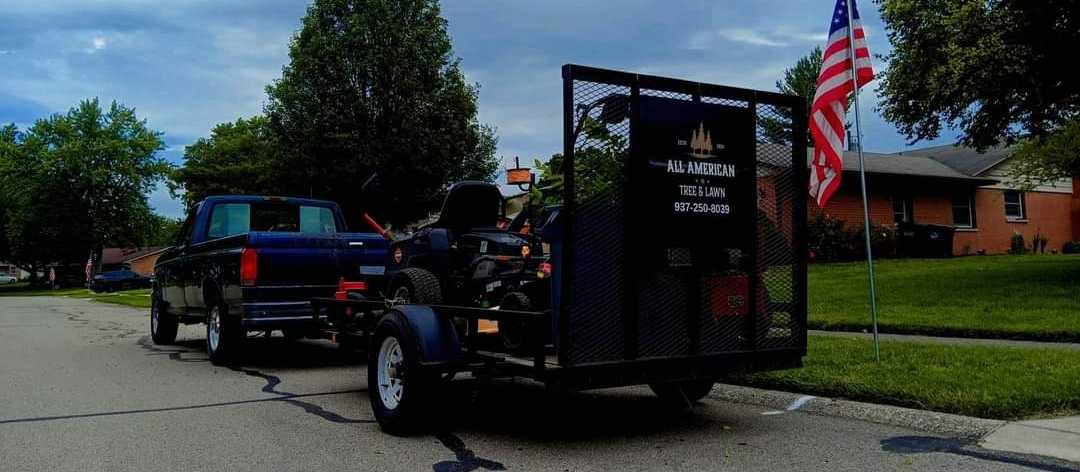 A black utility trailer with a mesh gate lettered All American Tree & Lawn and 937-250-8039, hitched to a pickup on a residential street beside an American flag.